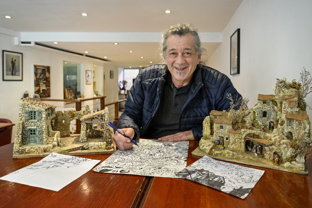 Francois Dalmas dessinant et posant à côté de l'une des crèches qu'il a créées sur une table du restaurant Le Borsalino - Agrandir l'image, fenêtre modale