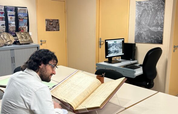 Un lecteur est assis devant un relevé nominatif de recensement de population et prend des notes sur un papier. Au fond, une borne informatique est allumée et montre une photo ancienne d'Aubagne