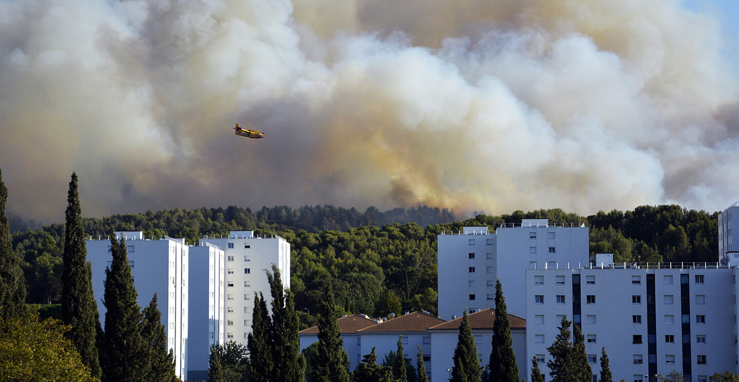 Vue du quartier du Charrel avec un feu de forêt en arrière-plan et un canadair qui survole la zone