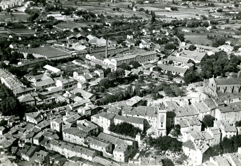 Photographie en noir et blanc. Vue aérienne du centre-ville d'Aubagne. Au premier plan, les maisons du centre ancien et la tour de l'horloge. Au second plan, l'ancien quartier des Défensions avec de grandes usines et des petites maisons avec jardin. A l'arrière-plan, la plaine cultivée de Beaudinard - Agrandir l'image, fenêtre modale