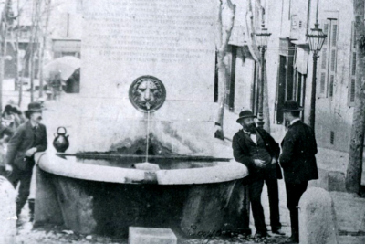 Trois hommes discutent près du monument. On aperçoit la partie basse du monument avec la plaque de marbre, le mascaron en forme de tête de lion et la vasque. - Agrandir l'image 1 sur 3, fenêtre modale
