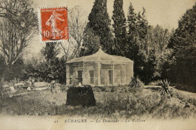 Au premier plan, une petite fontaine en rocaille devant une volière en forme de chapiteau entourée de jeunes palmiers. Trois personnes sont assises sur un banc à gauche de l'image. - Agrandir l'image 4 sur 9, fenêtre modale
