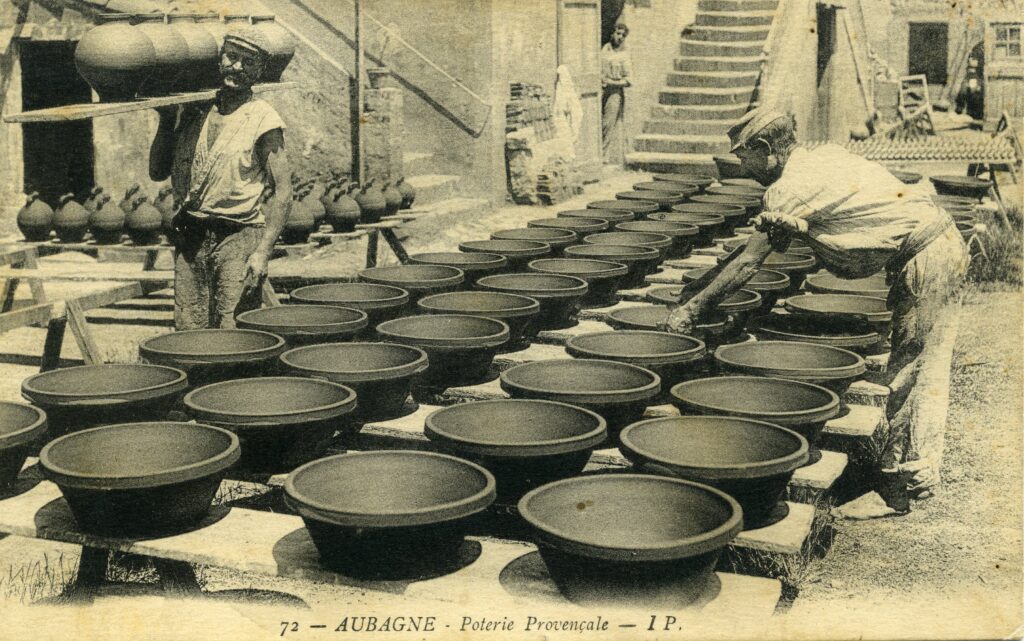 Un ouvrier de la poterie Ravel met à sécher un tian au milieu de trois files de tians, un autre porte plusieurs jarres sur une planche en bois. - Agrandir l'image, fenêtre modale