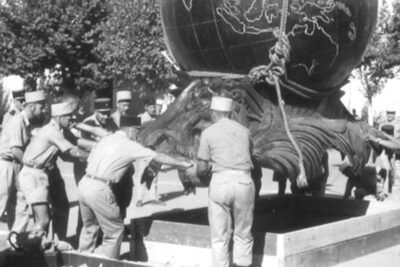 Des soldats aident à déplacer le Monument aux Morts (un gros globe terrestre) qui est tenu par des cordes et soulevé par une grue. - Agrandir l'image 1 sur 2, fenêtre modale