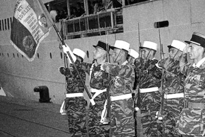 Quelques soldats sont au garde-à-vous devant un paquebot. Le premier d'entre eux brandit le drapeau du 1er RE. - Agrandir l'image 1 sur 2, fenêtre modale