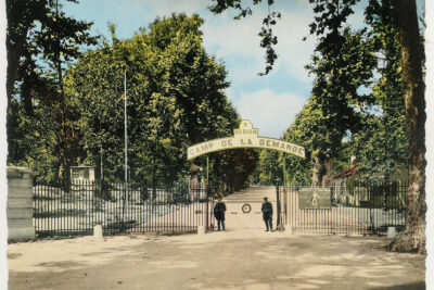 Grille d'entrée avec deux gendarmes en faction devant la barrière. Derrière la grille, on peut voir une longue allée bordée de platanes - Agrandir l'image 5 sur 9, fenêtre modale