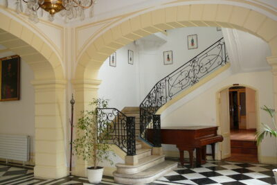 Montée d'escalier avec rampe en fer forgé noir et marches en marbre. Sous l'escalier, un piano marron. La montée d'escalier est ornée de portrait aquarellés de légionnaires en habits de différentes époques. Le sol est en carreaux noir et blanc - Agrandir l'image 9 sur 9, fenêtre modale