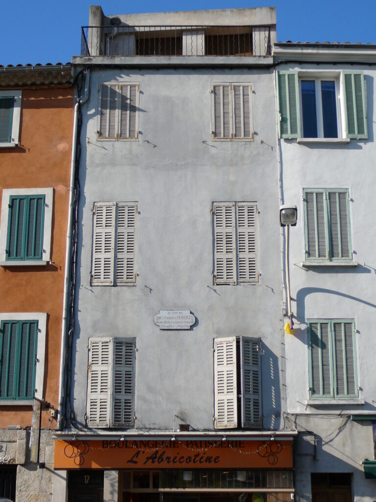 Immeuble gris étroit avec deux fenêtres à chacun des 3 étages et un balcon au quatrième. Une plaque murale est apposée sur le mur. Au rez-de-chaussée, le haut de la devanture d'une ancienne boulangerie appelée "L'Abricotine" - Agrandir l'image, fenêtre modale