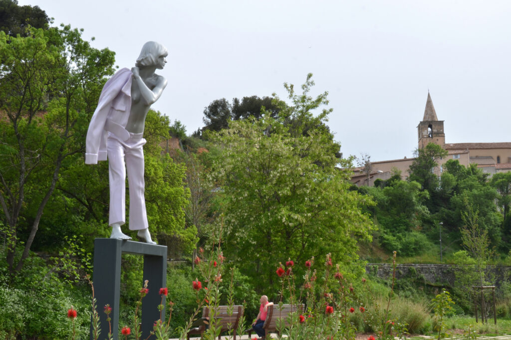 Statue d'une femme torse-nu en pantalon de costume en train d'enfiler une veste. Au fond, le clocher de l'église - Agrandir l'image, fenêtre modale