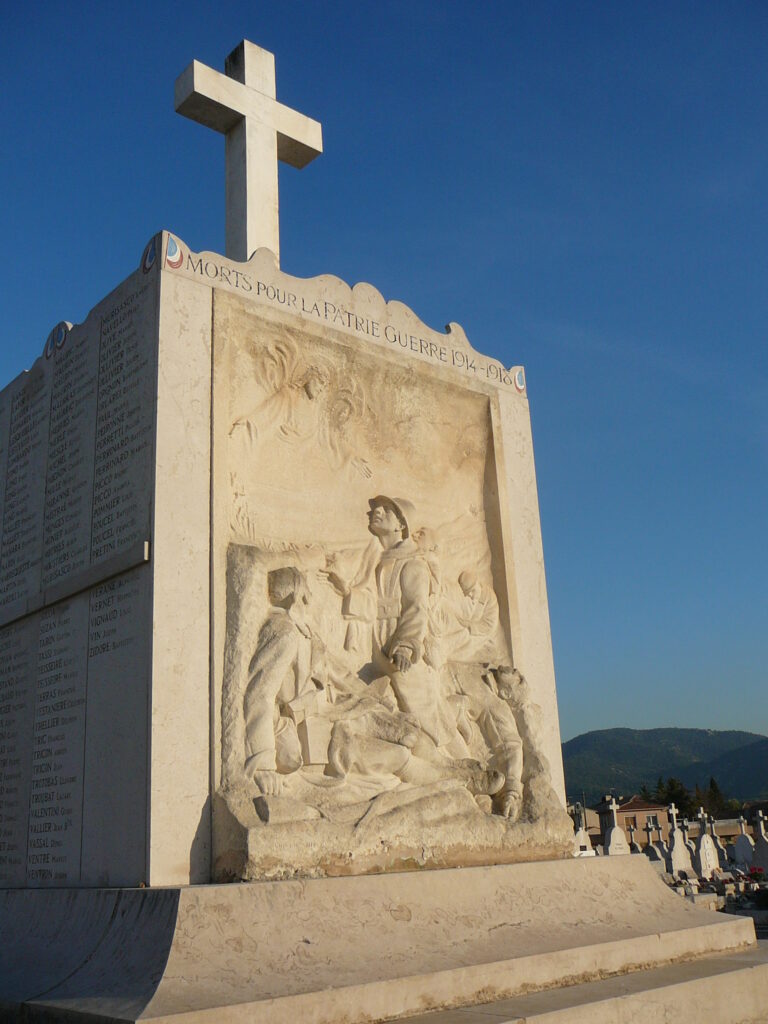 Le Monument aux Morts du cimetière des Passons illuminé par le soleil devant un ciel bleu. Sur le monument, un bas-relief de trois soldats dans une tranchée, deux regardant un ange, le troisième décédé - Agrandir l'image, fenêtre modale