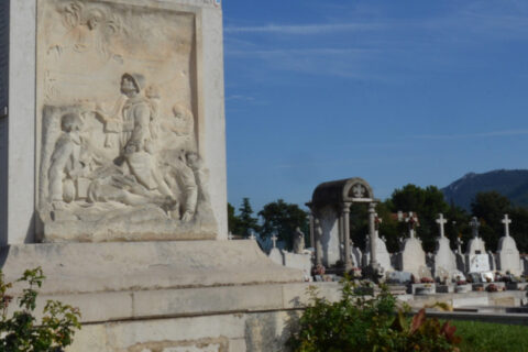Le Monument aux Morts du cimetière des Passons illuminé par le soleil devant un ciel bleu. Sur le monument, un bas-relief sculpté de trois soldats dont l'un est décédé et deux autres regardent un saint dans le ciel