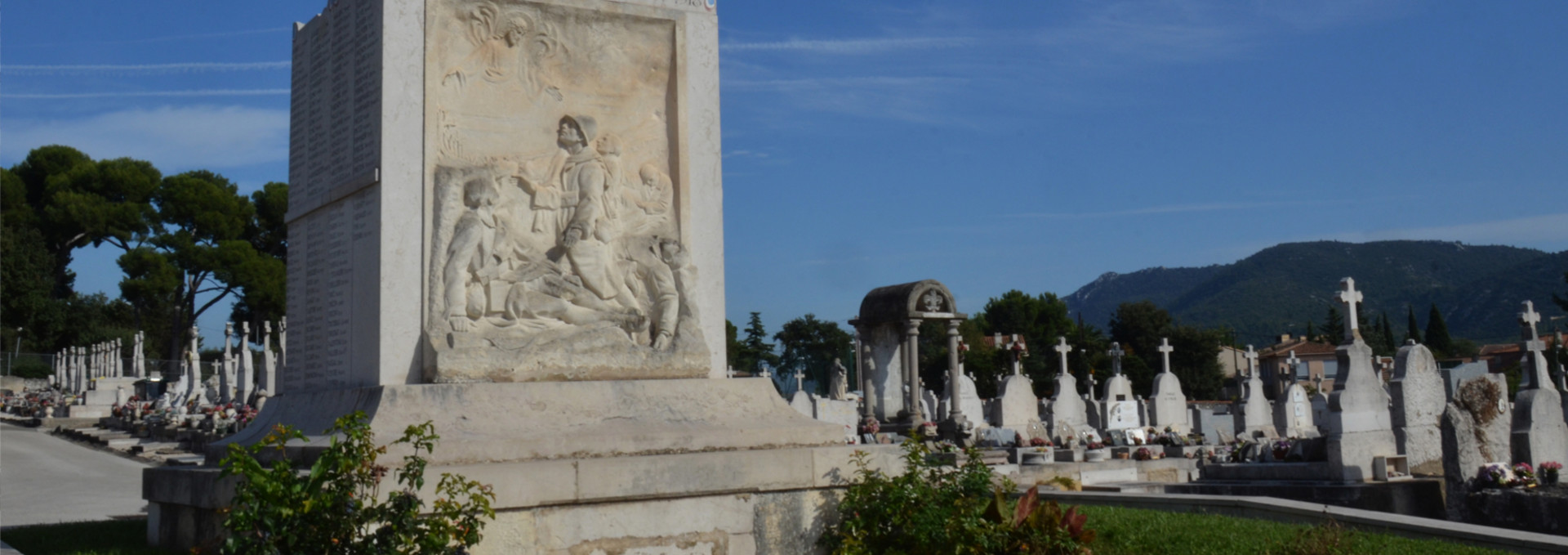 Le Monument aux Morts du cimetière des Passons illuminé par le soleil devant un ciel bleu. Sur le monument, un bas-relief sculpté de trois soldats dont l'un est décédé et deux autres regardent un saint dans le ciel