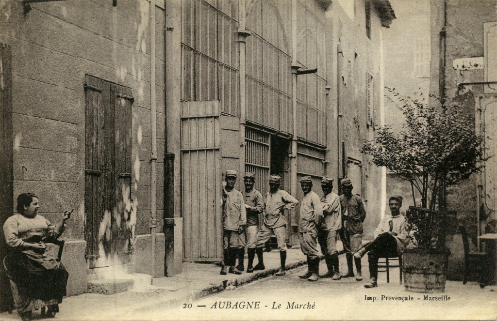 Des soldats posent dans la rue à l'arrière des halles. A gauche, une femme assise sur une chaise tricote et les montre du doigt - Agrandir l'image, fenêtre modale