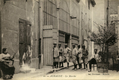 Des soldats posent dans la rue à l'arrière des halles. A gauche, une femme assise sur une chaise tricote et les montre du doigt - Agrandir l'image 1 sur 2, fenêtre modale