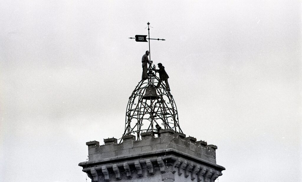 Deux hommes au sommet de la tour de l'horloge posent les décorations de Noël sur le campanile en fer forgé. On voit également la flèche de la tour avec l'inscription "1900" - Agrandir l'image, fenêtre modale