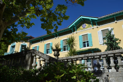 Au premier plan la balustrade de la terrasse, au second la façade de la bastide où l'on voit bien une frise en mosaïque jaune décorée de feuilles vertes et de roses, au centre un M stylisé dans un rond bleu. - Agrandir l'image 3 sur 6, fenêtre modale