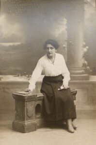 Photo en noir et blanc de Marie-Thérèse Castelin assise sur une rambarde de balcon devant un décor de colonne dorique et de fleurs - Agrandir l'image, fenêtre modale