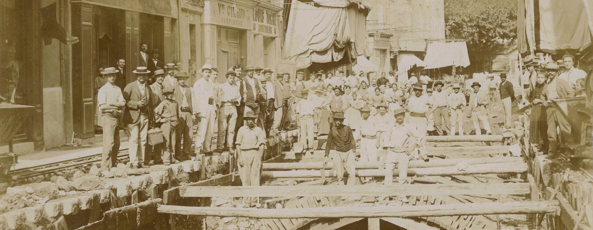 De nombreux ouvriers posent autour des travaux. On distingue la voûte qui couvre le Merlançon dans la rue de la République et on voit la rivière couler.