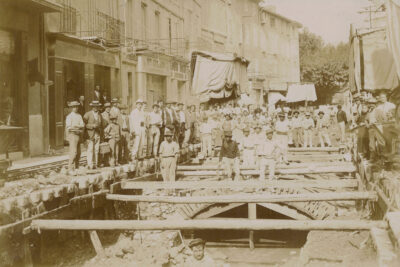 De nombreux ouvriers posent autour des travaux. On distingue la voûte qui couvre le Merlançon dans la rue de la République et on voit la rivière couler. - Agrandir l'image 7 sur 18, fenêtre modale