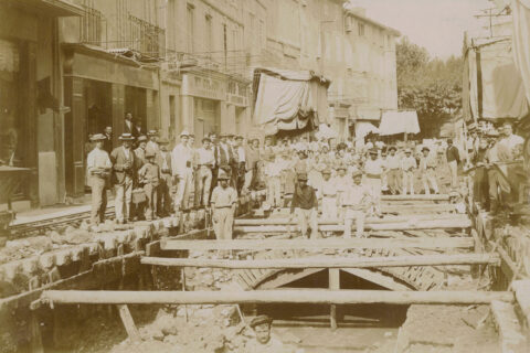 De nombreux ouvriers posent autour des travaux. On distingue la voûte qui couvre le Merlançon dans la rue de la République et on voit la rivière couler.