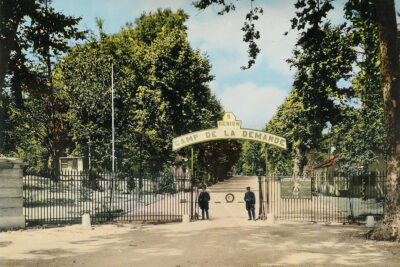 Photo en couleur. Des barrières et deux soldats en faction ferment l'entrée du camp. Une enseigne "9 région camp de la demande". Derrière les barrières, un chemin en terre traverse un espace très arboré. - Agrandir l'image 16 sur 18, fenêtre modale