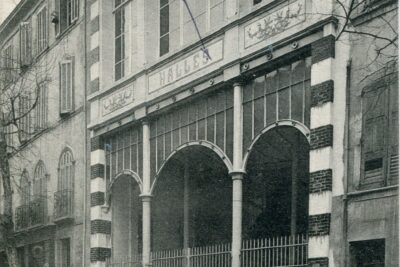 Au rez-de-chaussée du bâtiment fermé par des grilles, deux colonnes en métal soutiennent trois arcades également en métal. Le fronton est composé d'une frise composée de fleurs en céramique, de deux panneaux de céramique à motif floral entourant un troisième panneau en céramique où il est inscrit "Halles". A l'étage, trois grandes baies vitrées. - Agrandir l'image 5 sur 18, fenêtre modale