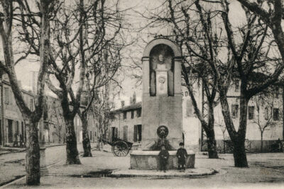 Fontaine entourée d'arbres sans feuilles et devant laquelle posent deux enfants. Au-dessus de la vasque, l'eau s'écoule depuis une tête de lion. Au sommet du monument, sous une voute soutenue par deux pilastres, le buste de l'abbé Barthélemy repose sur un socle. - Agrandir l'image 10 sur 18, fenêtre modale