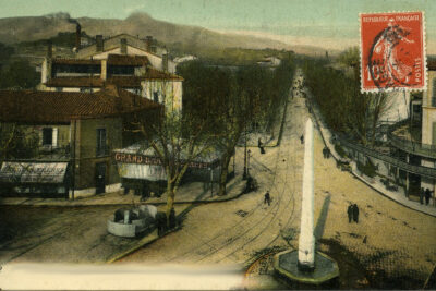 Photographie ancienne colorisée d'une vue aérienne de la place orientée vers le nord. Au premier plan, une fontaine octogonale surmontée d'un obélisque en pierre. La route pavée est creusée par des rails de tramway. De chaque côté, le grand bar des Glaces (à gauche) et le grand Café (à droite). Le cours Foch est masqué par les platanes sans feuilles. Au fond, le Garlaban. - Agrandir l'image 2 sur 18, fenêtre modale