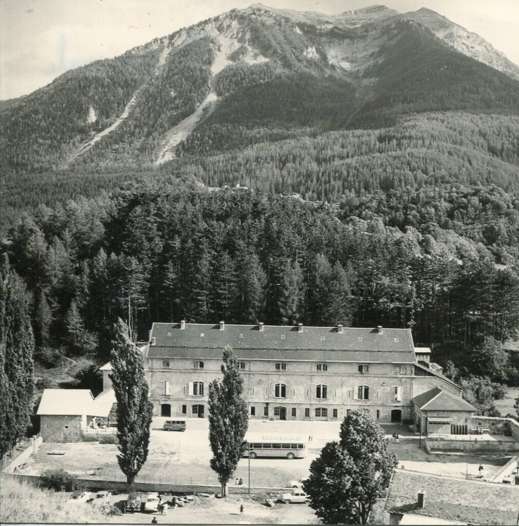 Vue en hauteur d'une grande bastide de 3 étages avec 7 fenêtres à chaque étage et une aile de chaque côté. Au fonds, la forêt et une haute montagne. - Agrandir l'image, fenêtre modale