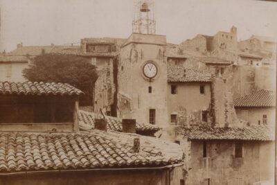 Photo sépia. Une tour sur laquelle figure un cadran surplombe des toitures. Les bâtiments autour semblent délabrés, certains portent les marques d'une démolition. Au sommet de la tour, un campanile en fer forgé abrite une petite cloche. - Agrandir l'image 3 sur 18, fenêtre modale
