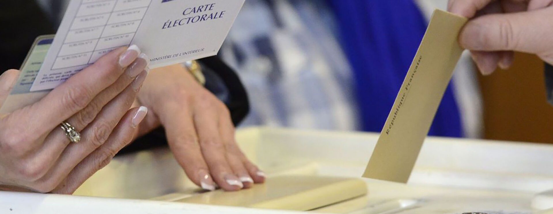 Une main tenant un bulletin de vote s'apprête à le glisser dans une urne, tandis qu'en face une autre main, qu'on devine être celle du président du bureau de vote, tient la carte d'électeur