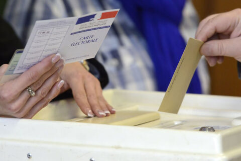 Une main tenant un bulletin de vote s'apprête à le glisser dans une urne, tandis qu'en face une autre main, qu'on devine être celle du président du bureau de vote, tient la carte d'électeur