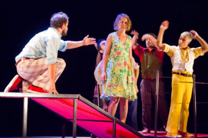 Photo d'une scène de la pièce "Angèle" par Cartoun Sardines - Plusieurs comédiens sont sur une scène avec des des habits aux couleurs chatoyantes, sur un fond noir - Agrandir l'image, fenêtre modale