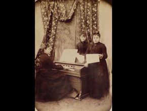 Augustine Camoin est assise sur une chaise et joue du piano devant deux femmes debout. Toutes les trois portent une robe noire - Agrandir l'image, .JPG 311,3 Ko (fenêtre modale)
